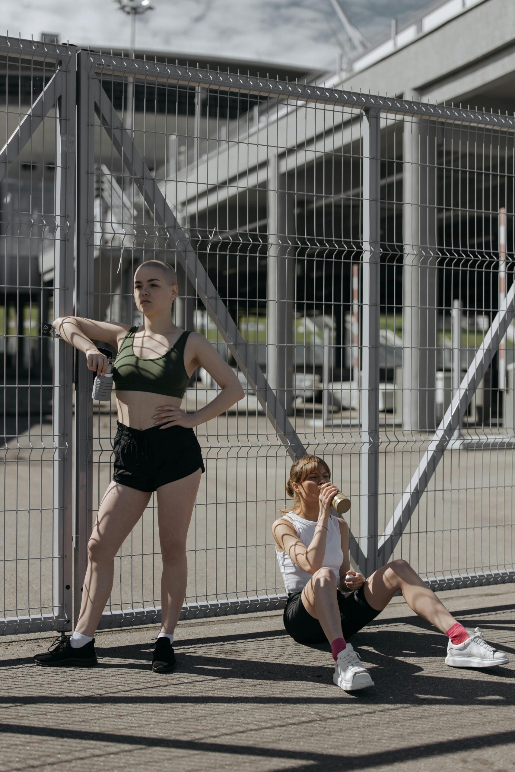 Two women in sportswear take a break by a fence in an urban outdoor setting, showcasing modern fitness lifestyle.